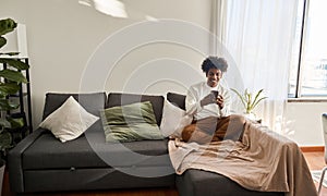 Smiling young African American guy sitting on couch using phone.