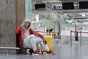 Smiling young African American hipster guy with suitcase calling on phone waiting flight in airport.