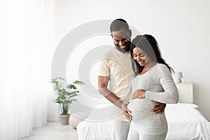 Smiling young african american husband hugs pregnant wife, touching belly in minimalist bedroom interior