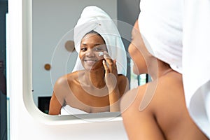 Smiling young african american woman applying face cream while looking in mirror, copy space