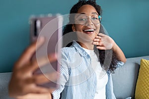 Smiling young african american woman using her mobile phone to take a selfie while sitting on sofa at home