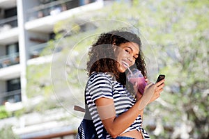 Smiling young african american woman walking with mobile phone and drinking smoothie