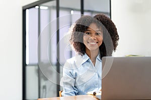 Smiling young African female entrepreneur sitting at a desk in her home office working online with a laptop