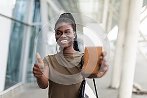 Smiling young african girl hold passport boarding pass tickets showing thumb up in airport terminal