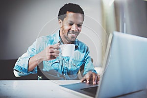 Smiling young African man making video conversation via laptop with partners while drinking white cup black tea in