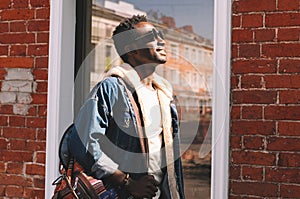 Smiling young african man wearing a jeans jacket and backpack looking up at sunlight while walking on a city street over