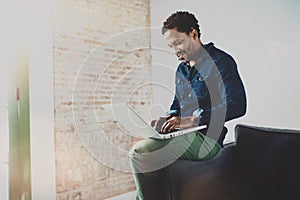 Smiling young African man working with laptop while sitting on sofa at his modern coworking place.Concept of happy