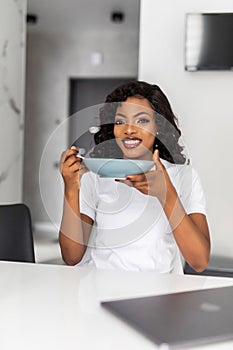 Smiling young african woman having breakfast while sitting at the kitchen with laptop