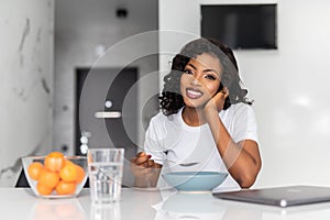 Smiling young african woman having breakfast while sitting at the kitchen with laptop