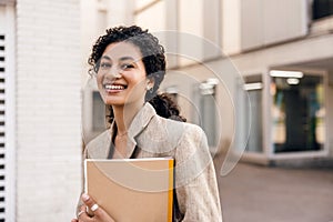 Smiling young african woman loking at camera, stands at street with folders.