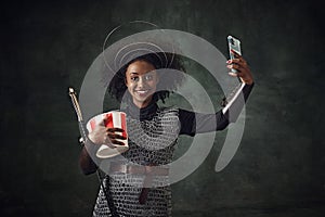 Smiling young African woman, medieval warrior in chainmail armor holding sword and taking selfie with mobile phone