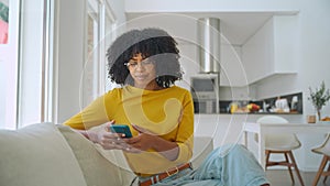 Smiling young African woman using cell phone relaxing on couch at home.