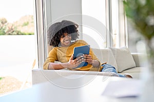 Smiling young African woman using digital tablet relaxing on couch at home.