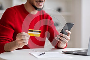 Smiling young arab guy with beard showing credit card and typing on smartphone in home office interior