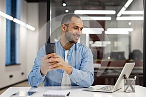 Smiling young arab man working in office on laptop, sitting at table and looking at screen, holding and using mobile