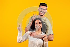 Smiling young arabic couple in casual hugging, showing peace sign with fingers and make horns, have fun