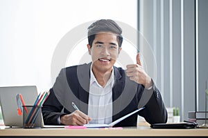 Smiling young asian business man working with note book on desk and show thumb up in a modern office