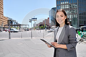 Smiling young Asian business woman standing on city street using digital tablet.
