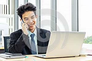 Smiling young asian businessman sitting at desk  talking on mobile phone and using laptop in workplace