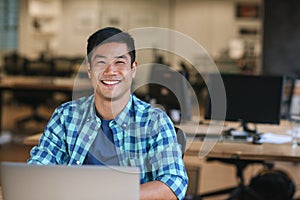 Smiling young Asian designer using a laptop at his desk