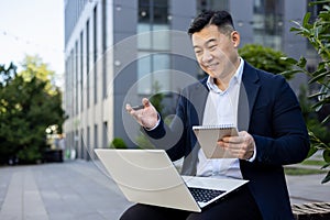 Smiling young Asian male businessman in a suit sitting outside an office building, holding a laptop on his lap, talking