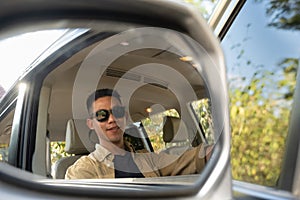 Smiling young Asian man looking throw the back view mirror while traveling by car in countryside