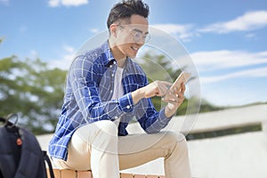 Smiling young asian man sitting on stairs and using smart phone