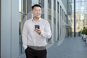 Smiling young Asian man walking near office center outside, holding phone in hand and looking away