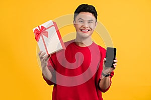 Smiling young Asian man wearing Christmas hat holding gift box and smartphone. Greeting and celebrating special day,  holiday