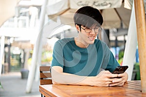 A smiling young Asian man wearing glasses sits at a wooden table outdoors, looking at his smartphone