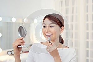 Smiling young Asian woman applying lipstick and looking to mirror