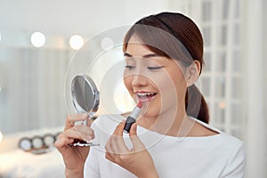 Smiling young Asian woman applying lipstick and looking to mirror