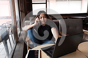 Smiling young Asian woman is calling with smartphone and online working with laptop in living room. Work from home for Covod-19