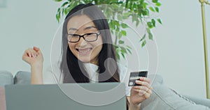 Smiling young Asian woman cheering after successfully making online payment using credit card from laptop at home