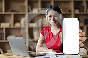 Smiling young asian woman with down syndrome in headphones with laptop shows phone with blank screen