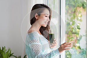 Smiling young asian woman drinking green fresh vegetable juice o