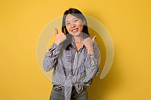 Smiling young Asian woman gesturing thumbs up with both hands, recommending something on yellow studio background
