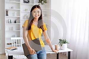 Smiling young asian woman holding closed laptop, standing in well-lit home office