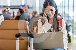 Smiling young asian woman sitting in waiting room and talking over smartphone with friend or family and using laptop. Beautiful