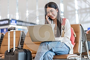 Smiling young asian woman sitting in waiting room and talking over smartphone with friend or family and using laptop. Beautiful
