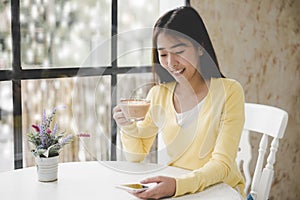 Smiling young asian woman sitting on windowsill and drinking tea at window in room. Good morning on winter day