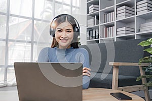 Smiling young asian woman using laptop web camera while wearing headphones and sitting on the rug beside to the sofa at