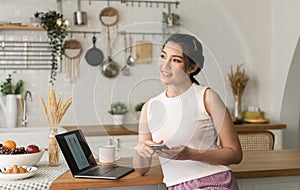 Smiling young asian woman using mobile phone while sitting in kitchen room at home with laptop computer