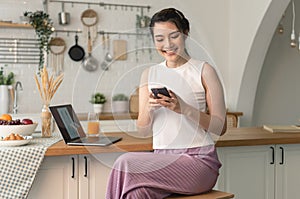Smiling young asian woman using mobile phone while sitting in kitchen room at home with laptop computer