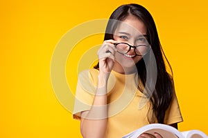 Smiling young asian woman wear eyeglasses holding book and looking at camera with smile face and happiness over yellow background,