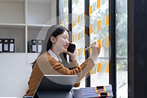 Smiling young asian woman writing on sticky notes on window in creative office