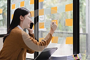 Smiling young asian woman writing on sticky notes on window in creative office