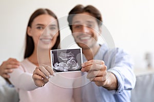 Smiling young attractive european wife and husband hold ultrasound scan picture unborn baby in room