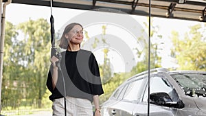 Smiling young attractive woman having fun with car washer gun, using like shot in camera. Girl is washing her vehicle at