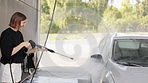 Smiling young attractive woman having fun with washing her vehicle at a self-service car wash, using a car cleaning tool